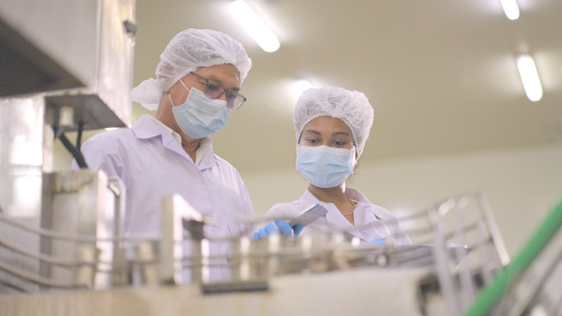 Product development team of industrial production is working form discuss the development quality of product according to the production standards. Two food factory employees in white sterile uniforms, discuss about product quantity or raw materials for production line in processed food factory or manufacturing industry.